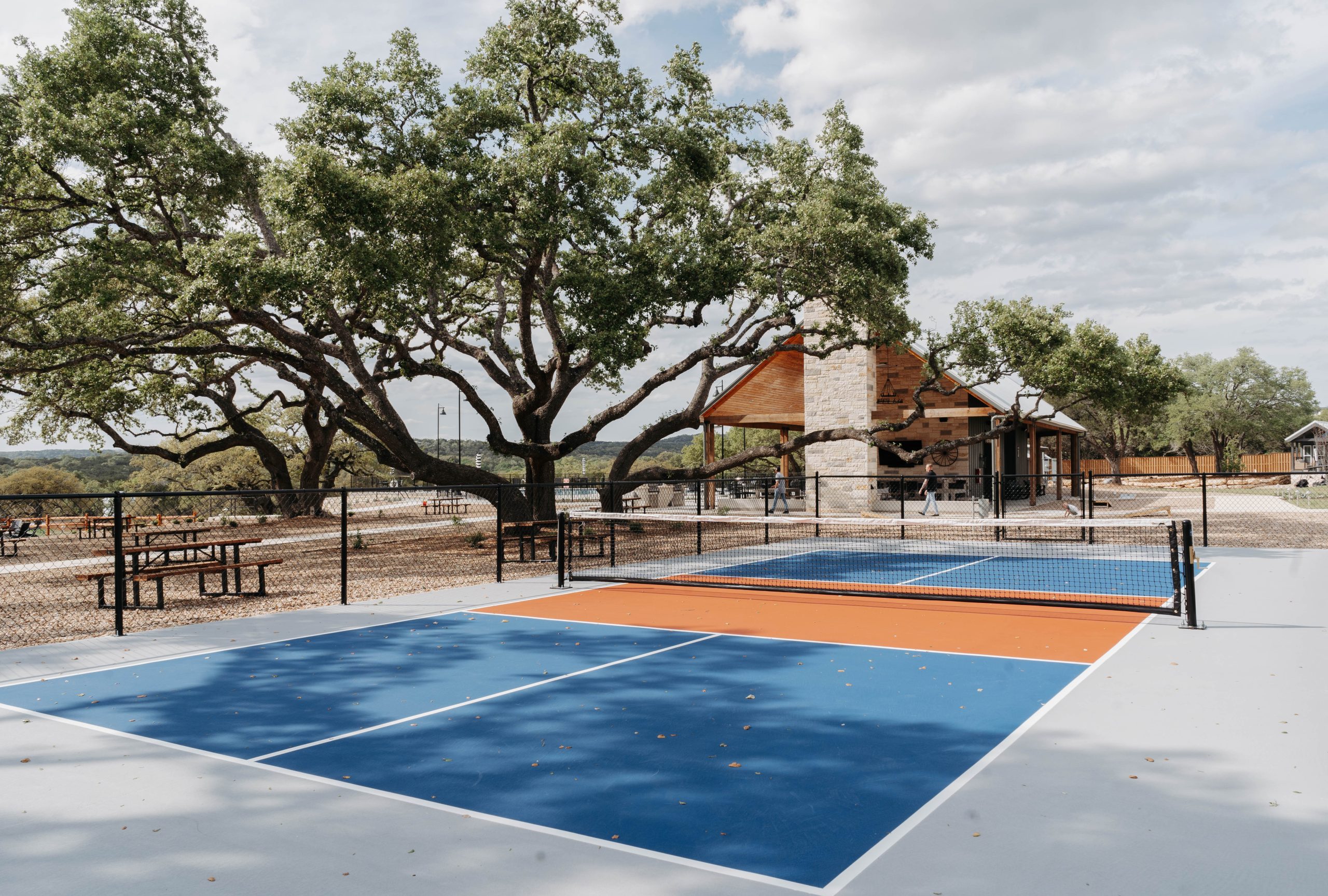 Pickleball courts shaded by a large oak with the Hill Country pavilion behind at Horseshoe Ridge RV Resort