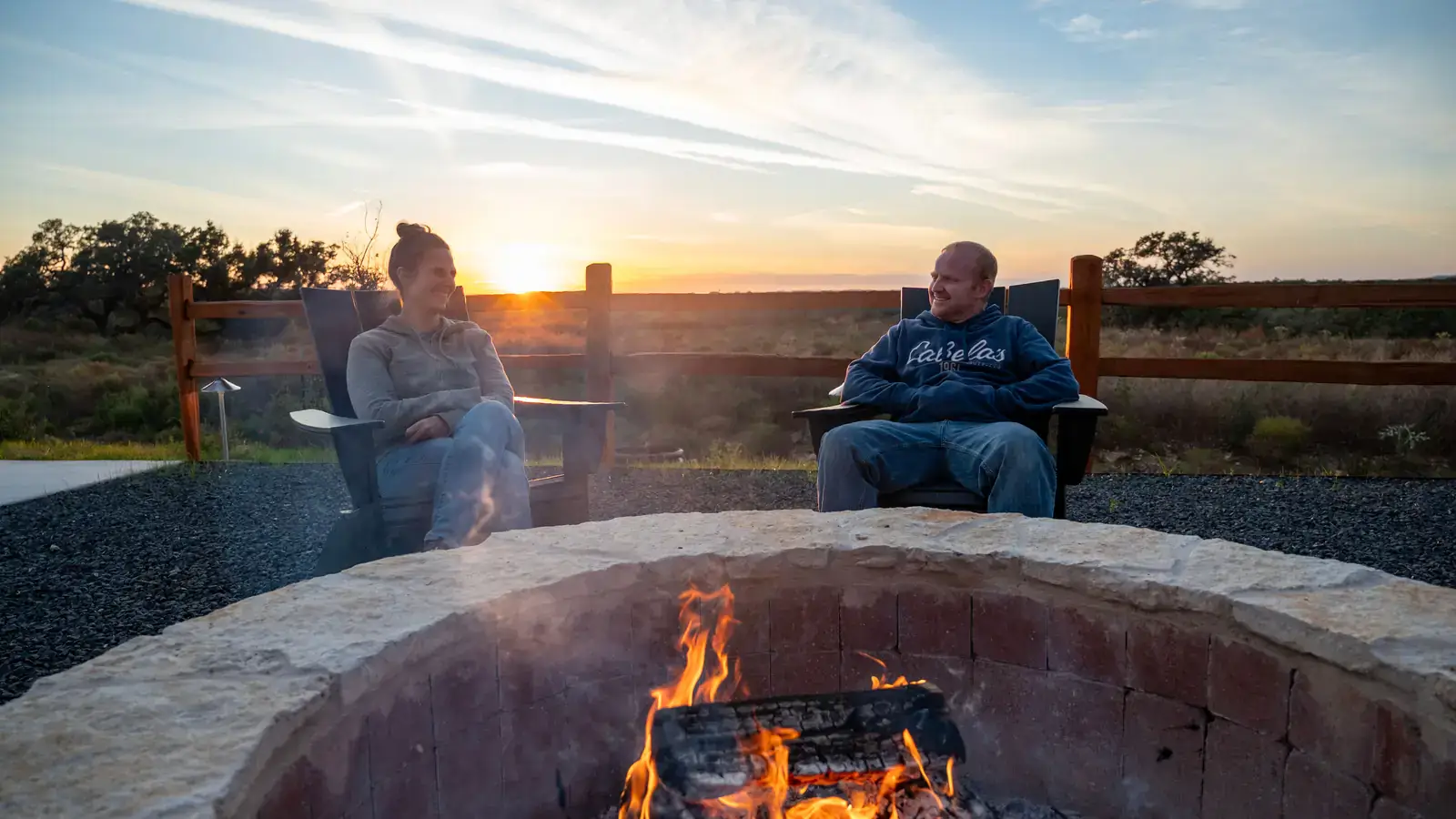 Couple relaxing by a stone fire pit at sunset, with Hill Country views beyond the fence
