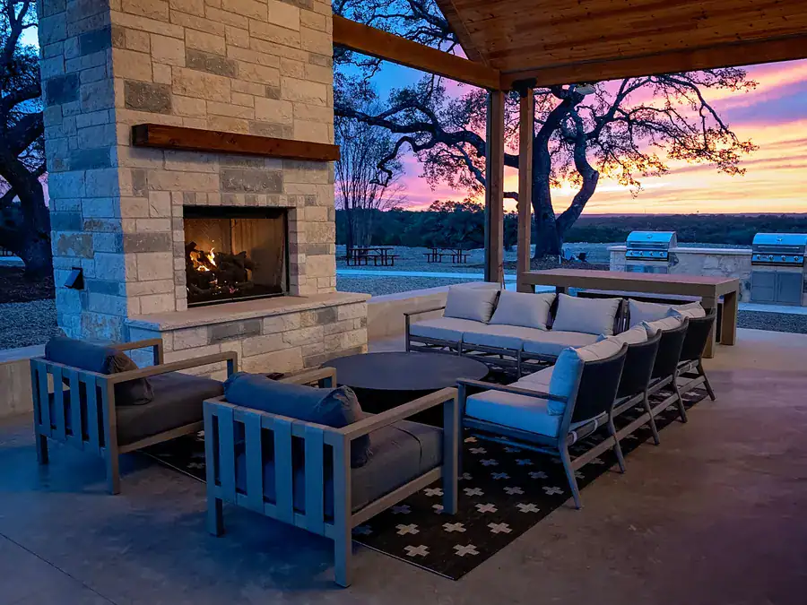 Covered pavilion with a lit stone fireplace, outdoor sofas and grills at dusk