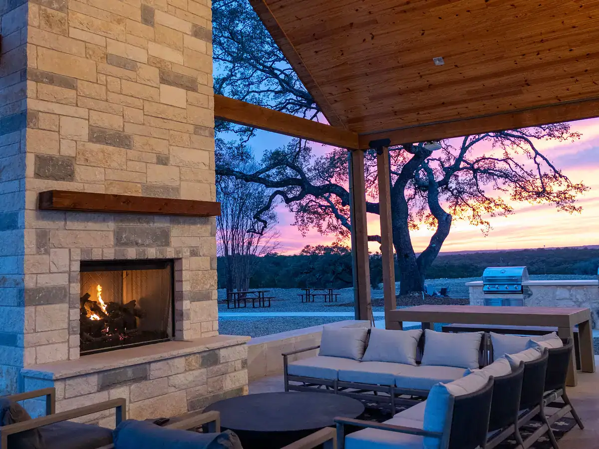The covered pavilion's stone fireplace lit at dusk, with outdoor sofas and oaks beyond