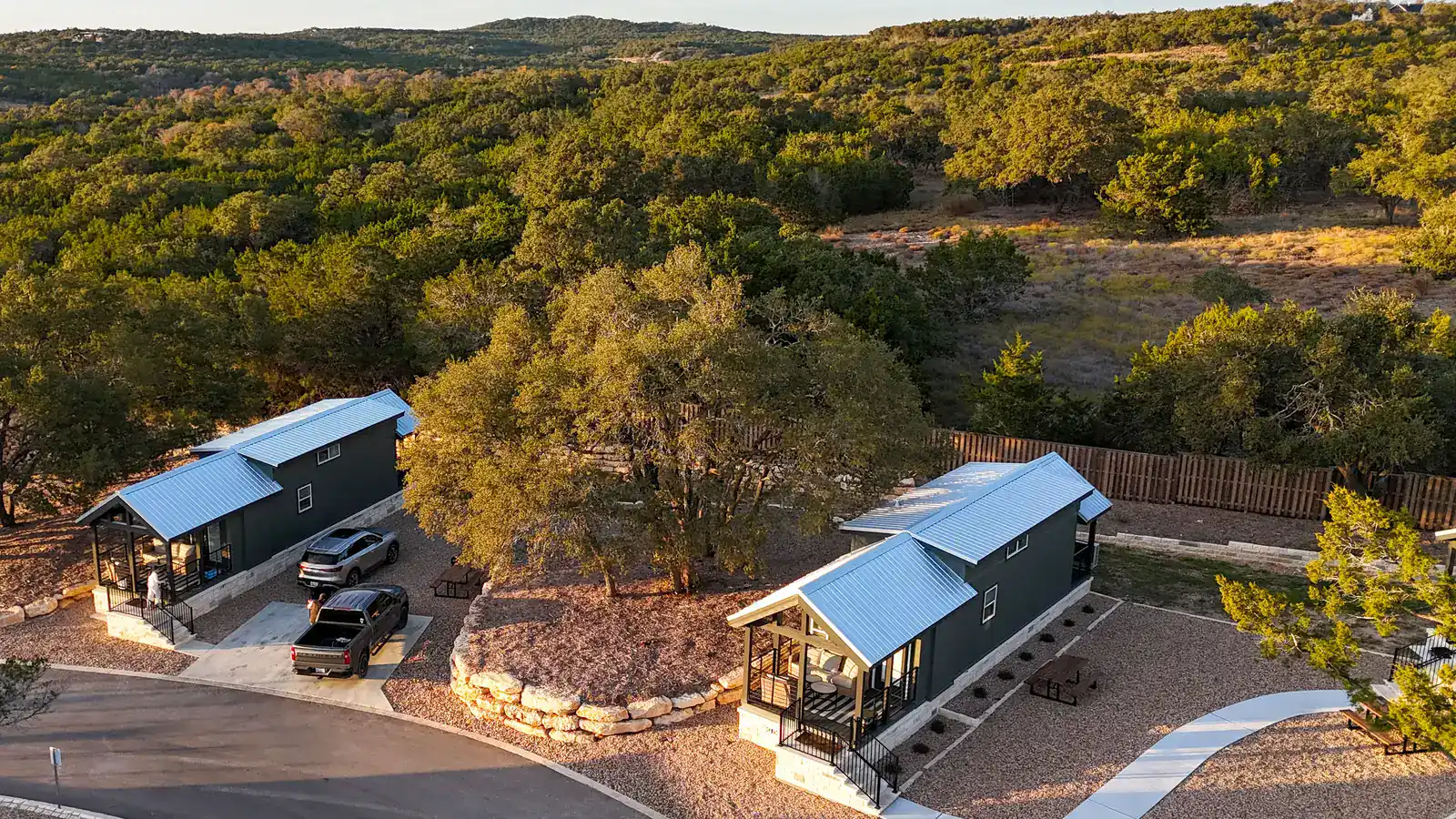 Golden-hour aerial of a Horseshoe Ridge cabin and its covered porch among the oaks, with the Texas Hill Country beyond